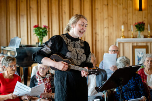Charlitte Stav spiller ukulele. I bakgrunnen ser vi kormedlemmene fra Hølonda Helsekor.