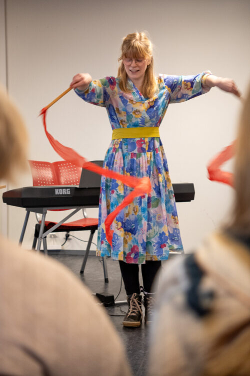 Kvinne står med en pinne i hver hånd som har et rødt silkebånd knyttet fast i seg. hun beveger pinnene slik at silketråden lager en spiral form. Foto: Alexandra Dahlen